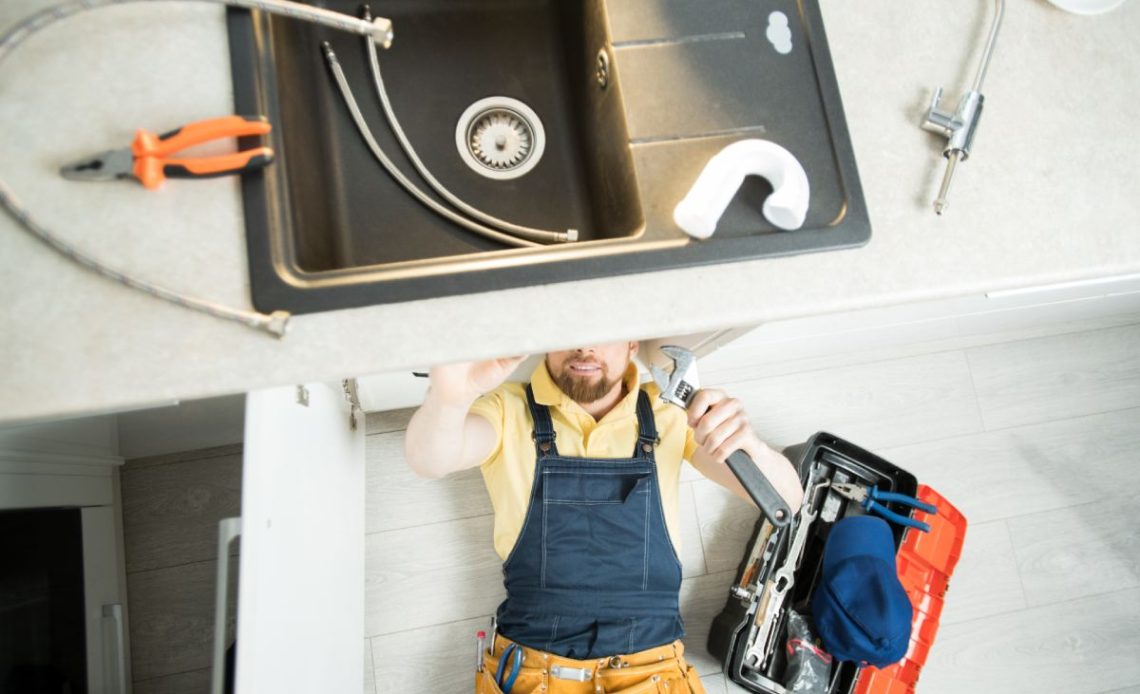 Directly above view of smiling handsome young plumber with beard lying on floor and using wrench while repairing water pipe in kitchen