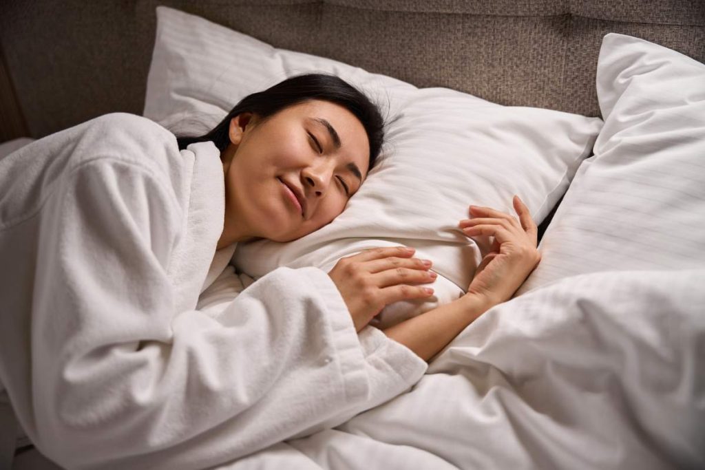 Pretty Asian woman in white soft bathrobe smiling in her sleep on comfortable orthopedic bed with fresh linen, enjoying sweet dreams and resting