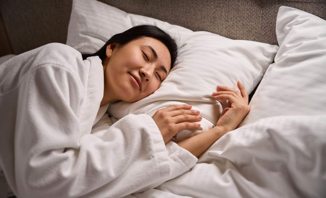 Pretty Asian woman in white soft bathrobe smiling in her sleep on comfortable orthopedic bed with fresh linen, enjoying sweet dreams and resting