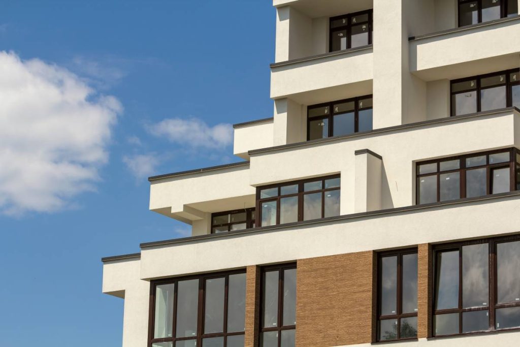 Close-up detail of new white apartment buildings with terraced balconies, shiny windows and low protective fence on flat roof top on blue sky background. Modern architecture, professional building.